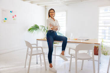 Erfolgreiche Geschäftsfrau, die in die Kamera lächelt, während sie auf einem Tisch in einem Sitzungssaal sitzt. Glückliche Unternehmerin, die darauf wartet, eine Sitzung an einem kreativen Arbeitsplatz zu beginnen. - JLPSF07350