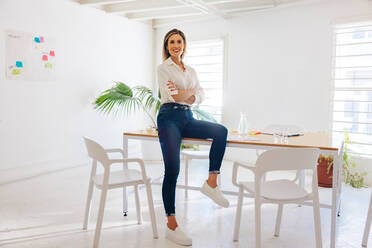 Successful businesswoman smiling at the camera while sitting on a table in a boardroom. Happy female entrepreneur waiting to start a meeting in a creative workplace. - JLPSF07350