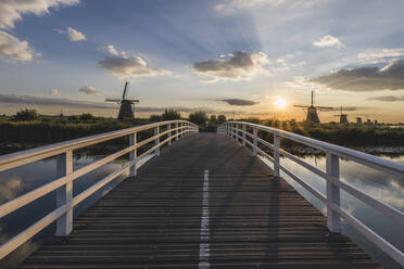 Niederlande, Südholland, Kinderdijk, Brücke auf dem Lande bei Sonnenuntergang mit historischen Windmühlen im Hintergrund - KEBF02415