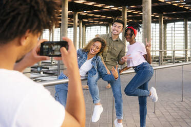 Junger Mann fotografiert Freunde mit dem Smartphone am Bahnhof - MMPF00358