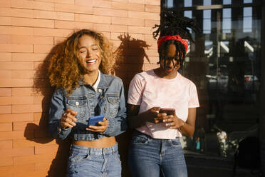 Young woman laughing by friend using mobile phone on sunny day - MMPF00367