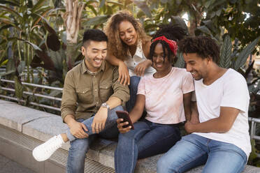 Lächelnde multirassische Männer und Frauen, die gemeinsam ein Smartphone an einer Wand benutzen - MMPF00400