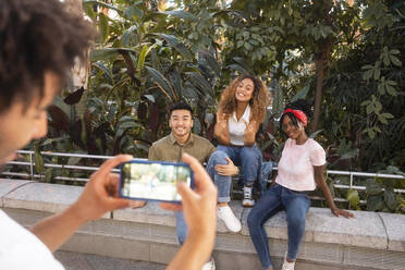 Young man photographing smiling friends through smart phone on wall - MMPF00402