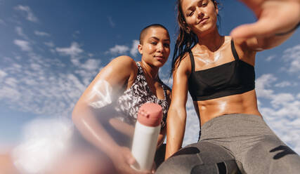 Two female friends sitting outdoors looking at camera with sun flare. Fitness women resting after workout on a wall. Pov shot. - JLPSF07366