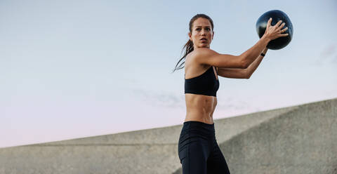 Athletic woman doing fitness training holding a medicine ball. Woman in fitness wear doing workout with a medicine ball outdoors. - JLPSF07368