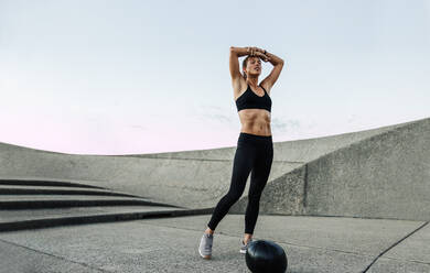 Female in sportswear feeling exhausted after intense training. Woman taking a break after workout outdoors with a medicine ball on ground. - JLPSF07369