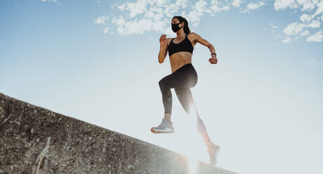 Niedrige Winkelaufnahme einer Fitness-Frau mit Gesichtsmaske, die über eine Stützmauer läuft. Eine Sportlerin trainiert an einem sonnigen Morgen. - JLPSF07400