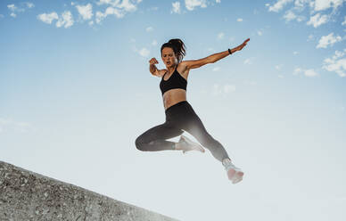 Fitness-Frau springt beim Training von der Wand. Niedriger Blickwinkel einer Sportlerin in Sportkleidung beim Training im Freien am Morgen. - JLPSF07403