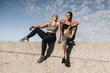 Two female friends sitting outdoors on a wall talking and smiling. Fitness women resting after workout on a wall. - JLPSF07413