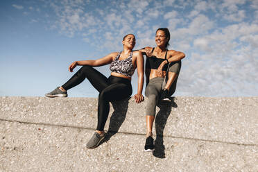 Zwei Freundinnen sitzen im Freien auf einer Mauer und reden und lächeln. Fitness-Frauen ruhen sich nach dem Training auf einer Mauer aus. - JLPSF07413