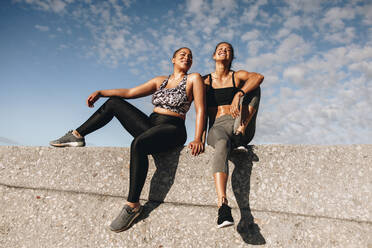 Zwei Fitness-Frauen entspannen sich nach dem Training im Freien. Freundinnen sitzen auf einer Mauer im Freien und lächeln. - JLPSF07414