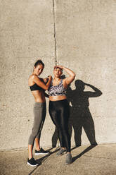 Frau und ihr Fitnesstrainer stehen an einem sonnigen Morgen im Freien an einer Wand. Frauen in Fitnesskleidung stehen zusammen und schauen in die Kamera. - JLPSF07418