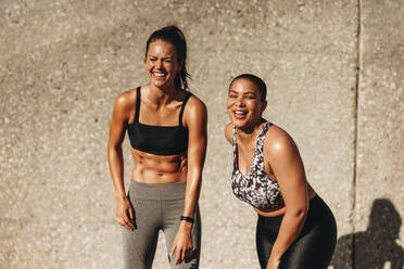 Zwei Fitness-Frauen, die morgens im Freien stehen. Fitness-Frauen in Trainingskleidung, die gemeinsam gegen eine Wand lächeln. - JLPSF07419