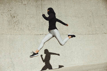 Woman wearing hijab training wear and protective face mask exercising outdoors. Woman running and jumping in sportswear. - JLPSF07435