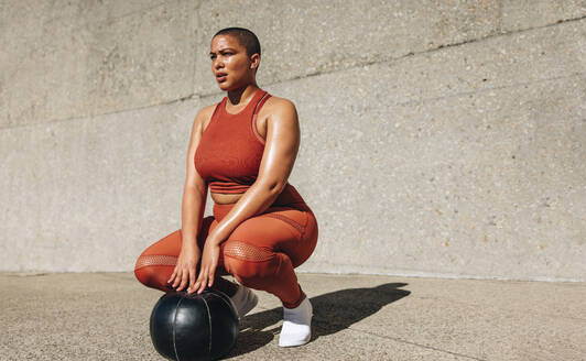 Fitness Frau hockt mit Medizinball auf dem Boden. Plus size Frau in Sportkleidung mit Medizinball schaut weg. - JLPSF07446