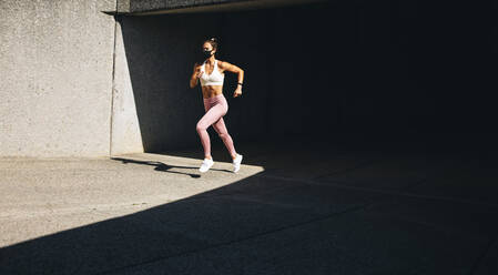 Ganzkörperaufnahme einer Frau beim morgendlichen Joggen mit Gesichtsmaske. Frau in Sportkleidung und mit Gesichtsmaske bei einer körperlichen Trainingseinheit. - JLPSF07461