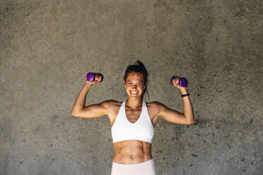 Smiling woman exercising with hand weights. Woman holding dumbbell flexing her biceps looking at camera and smiling against a wall. - JLPSF07473