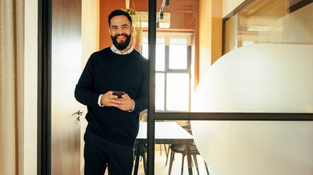 Successful young businessman smiling while holding a smartphone in a modern office. Cheerful young businessman sending a text message while standing in a co-working space. - JLPSF07486