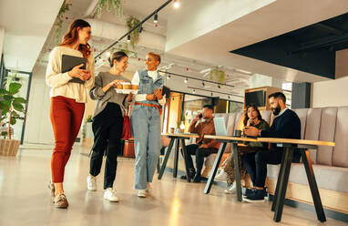 Lächelnde Geschäftsfrauen, die morgens durch ein modernes Büro gehen. Eine Gruppe junger Geschäftsfrauen, die sich fröhlich unterhalten. Glückliche Unternehmerinnen, die in einem Co-Working Space zusammenarbeiten. - JLPSF07492