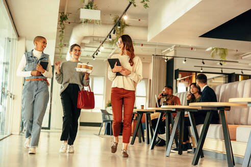 Drei glückliche Geschäftsfrauen gehen morgens durch ein modernes Büro. Eine Gruppe junger Geschäftsfrauen unterhält sich fröhlich. Unternehmerinnen arbeiten in einem Co-Working Space zusammen. - JLPSF07493