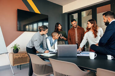 Verschiedene Geschäftsleute besprechen einige Berichte in einem Sitzungssaal. Eine Gruppe multikultureller Geschäftsleute steht in einem modernen Büro um einen Tisch herum. Junge Unternehmer arbeiten gemeinsam an einem Projekt. - JLPSF07523