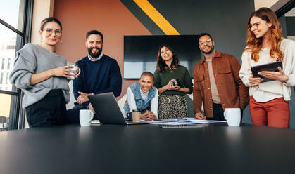 Junge Geschäftsleute, die in einem Sitzungssaal fröhlich lächeln. Eine Gruppe multikultureller Geschäftsleute, die in einem modernen Büro um einen Tisch herumstehen. Erfolgreiche junge Unternehmer, die als Team zusammenarbeiten. - JLPSF07526