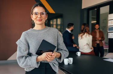 Modern businesswoman looking at the camera while holding a digital tablet. Cheerful young businesswoman standing in a boardroom with her colleagues in the background. - JLPSF07532