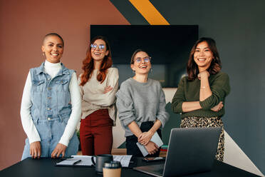 Erfolgreiche Unternehmerinnen, die fröhlich lächelnd hinter einem Tisch in einem Sitzungssaal stehen. Gruppe von multikulturellen Geschäftsfrauen, die in einem modernen Büro zusammenarbeiten. - JLPSF07536