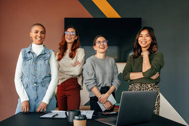 Successful female entrepreneurs smiling cheerfully while standing behind a table in a boardroom. Group of multicultural businesswomen working together in a modern office. - JLPSF07536