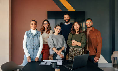 Successful business team smiling at the camera while standing behind a table in a boardroom. Group of multicultural businesspeople working together in a modern workplace. - JLPSF07537