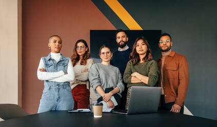 Eine Gruppe multikultureller Unternehmer, die an einem modernen Arbeitsplatz zusammenarbeiten, blickt in die Kamera, während sie hinter einem Tisch in einem Sitzungssaal stehen. - JLPSF07538