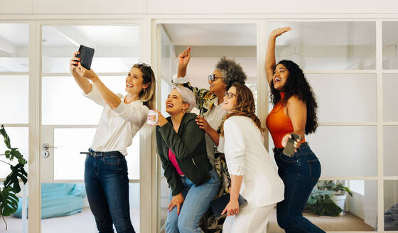 Eine Gruppe fröhlicher Geschäftsleute lächelt glücklich, während sie in einem modernen Büro ein gemeinsames Selfie machen. Multikulturelle Geschäftsleute feiern ihren Erfolg als Team. - JLPSF07597
