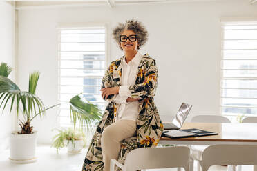 Happy businesswoman smiling at the camera while sitting on a table in a meeting room. Mature businesswoman waiting for a meeting in a modern office. - JLPSF07598