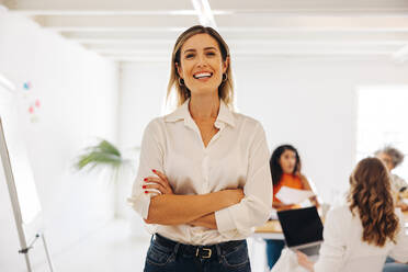 Glückliche junge Geschäftsfrau, die in die Kamera lächelt, während sie in einem Besprechungsraum steht. Fröhliche junge Geschäftsfrau, die an einer Besprechung mit ihren Kollegen in einem reinen Frauenbüro teilnimmt. - JLPSF07606