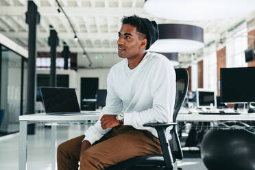 Young businessman looking away while sitting in an office. Creative businessman taking a break while working on a new project in a modern workplace. Young software developer sitting alone. - JLPSF07629