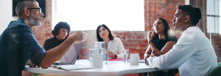 Manager leading a meeting in a modern workplace. Experienced mature businessman briefing his colleagues in a meeting room. Group of creative businesspeople working as a team. - JLPSF07635