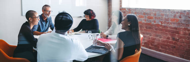 Lächelnde Geschäftsleute, die in einem Besprechungsraum zusammensitzen. Eine Gruppe erfolgreicher Geschäftsleute, die an ihrer morgendlichen Besprechung in einem modernen Büro teilnehmen. Gut gelaunte Kollegen, die zusammenarbeiten. - JLPSF07637