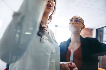 Selbstbewusste Geschäftsfrauen beim Brainstorming in einem kreativen Büro. Zwei konzentrierte junge Geschäftsfrauen kleben Haftnotizen an eine Glaswand und tauschen ihre Ideen an einem modernen Arbeitsplatz aus. - JLPSF07648