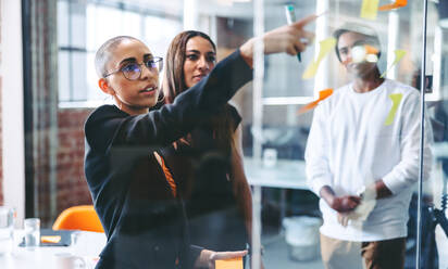 Smart businesswoman sharing her ideas with her team. Confident young businesswoman sharing her sticky notes with her colleagues while standing in front of a glass wall. Businesspeople working together. - JLPSF07649