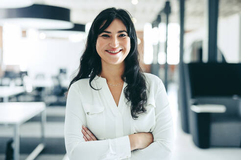 Successful businesswoman smiling at the camera. Happy young businesswoman standing with her arms crossed in a modern workplace. Cheerful businesswoman standing alone in an office. - JLPSF07653