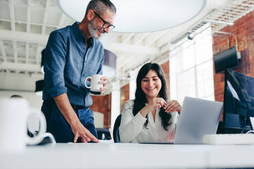 Two happy colleagues collaborating on a new project in the office. Mature businessman having a discussion with his female colleague while standing at her desk in a modern workplace. - JLPSF07660