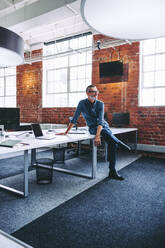 Happy mature businessman sitting on an office desk. Mature businessman smiling while sitting alone in a modern office. Experienced businessman feeling content with his success in the office. - JLPSF07665