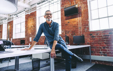 Mature businessman smiling while sitting in a modern workplace. Happy mature businessman sitting alone on an office desk. Experienced businessman feeling content with his success in the office. - JLPSF07666