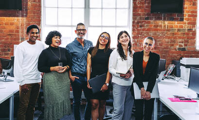 Successful businesspeople smiling cheerfully in an office. Group of happy businesspeople standing together in a creative workplace. Business colleagues enjoying working together. - JLPSF07669