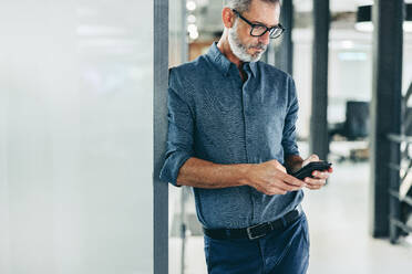 Kreativer Geschäftsmann, der eine Textnachricht in einem Büro sendet. Reifer Geschäftsmann, der ein Smartphone benutzt, um mit seinen Kunden zu kommunizieren. Erfahrener Geschäftsmann, der allein an einem modernen Arbeitsplatz steht. - JLPSF07672