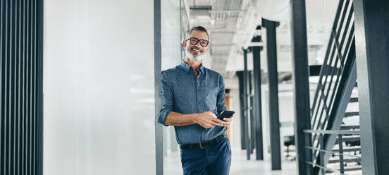 Smiling mature businessman holding a smartphone in an office. Happy businessman looking at the camera while standing alone in a modern workplace. Experienced businessman communicating with his clients. - JLPSF07673