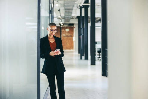 Selbstbewusste Geschäftsfrau, die in einem kreativen Büro eine Textnachricht sendet. Junge Geschäftsfrau, die mit ihren Kunden über ein Smartphone kommuniziert. Geschäftsfrau, die an einem modernen Arbeitsplatz steht. - JLPSF07676