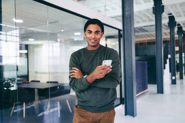 Creative young businessman holding a smartphone in a modern office. Happy young businessman looking at the camera while standing alone in a workplace. Young businessman sending a text message. - JLPSF07681