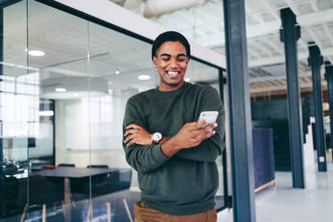 Gute Nachrichten im Büro. Glücklicher junger Geschäftsmann, der fröhlich lächelt, während er eine Textnachricht in einem modernen Büro liest. Erfolgreicher Geschäftsmann, der ein Smartphone hält, während er an einem kreativen Arbeitsplatz steht. - JLPSF07682