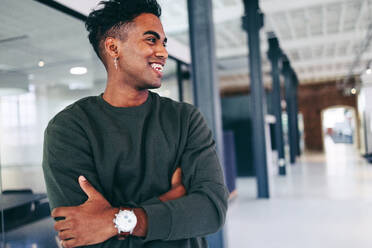 Modern businessman smiling while standing alone in a creative office. Happy young businessman looking away cheerfully while standing with his arms crossed. Businessman wearing business casual clothing. - JLPSF07683